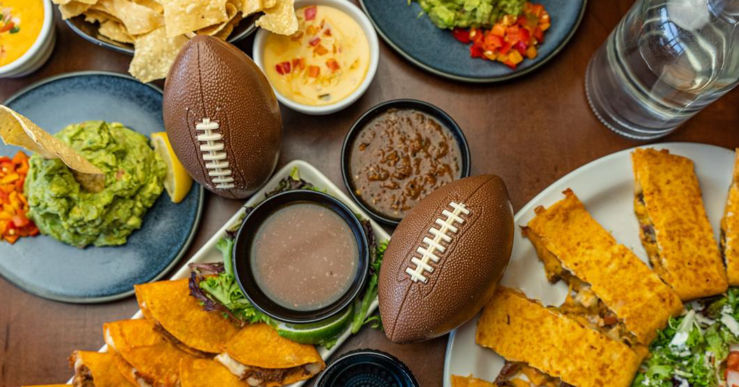 A table filled with Mexican appetizers like guacamole, quesadillas, tacos and queso paired with football decorations